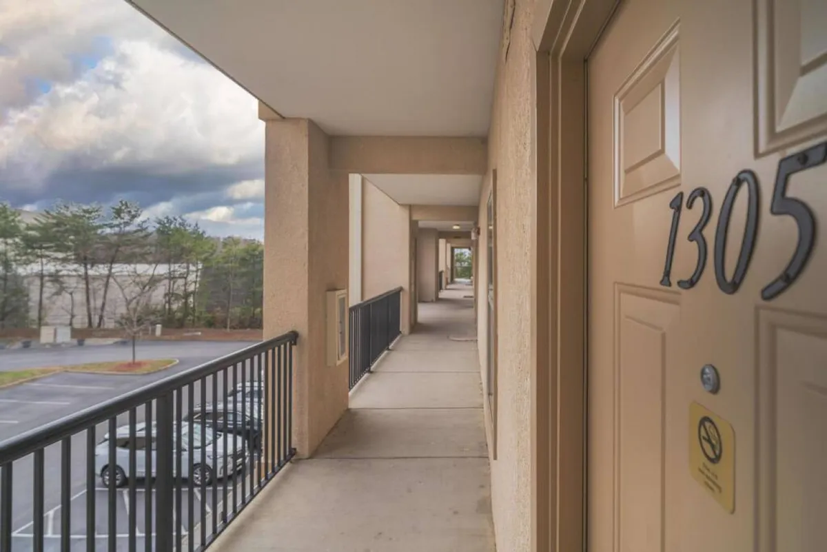 A view from a unit's exterior corridor, showing the door number 1305, a railing, and a glimpse of the parking lot with trees in the background. The corridor appears flat and accessible, suitable for wheelchair users or guests with mobility needs.