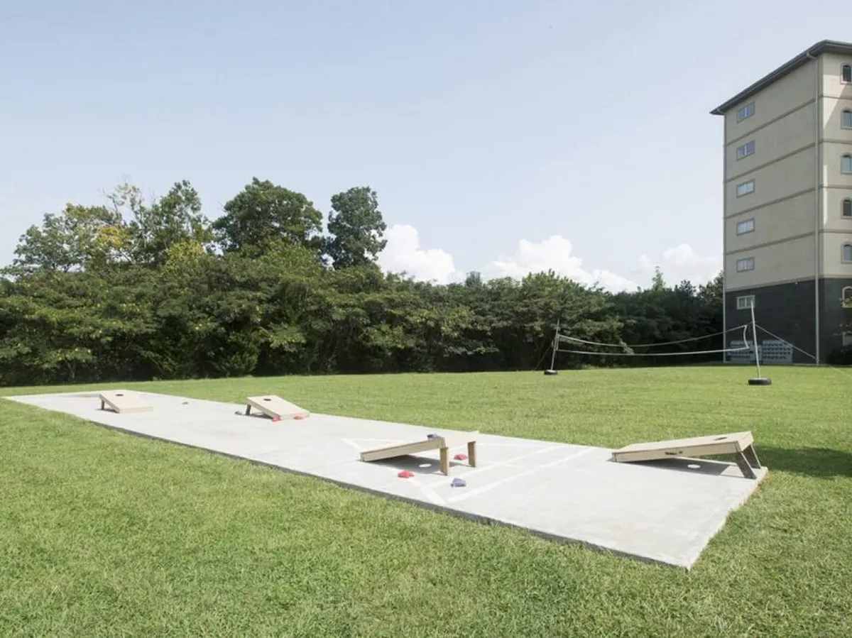 A vibrant outdoor scene featuring cornhole boards and a volleyball net set up on a grassy area, with a building in the background. The image conveys an atmosphere of friendly competition and outdoor play, inviting families to engage in games and social activities.