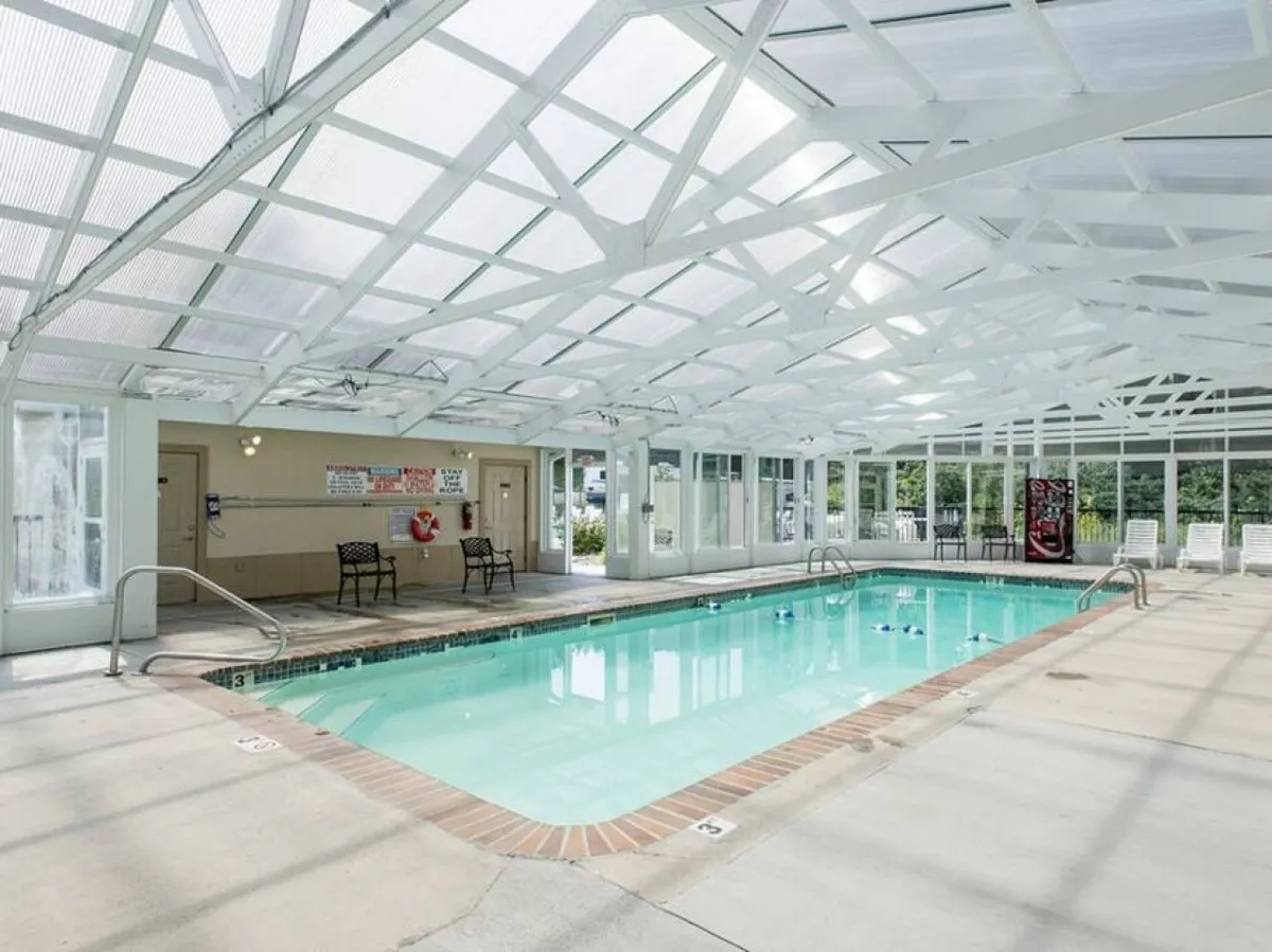 This image showcases a bright, spacious indoor swimming pool surrounded by chairs, with a transparent roof that allows natural sunlight to stream in. The inviting pool is perfect for year-round swimming and family play, while the ample seating encourages socializing and relaxation.