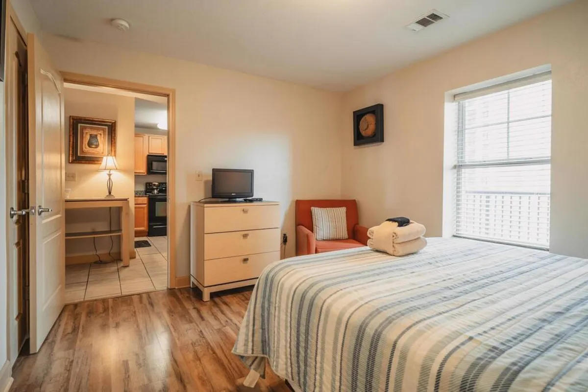 A warm, welcoming bedroom featuring a bed with a striped blanket, a comfortable orange chair, and a television atop a dresser. The space exudes a peaceful, lived-in feel ideal for relaxation or quiet time.
