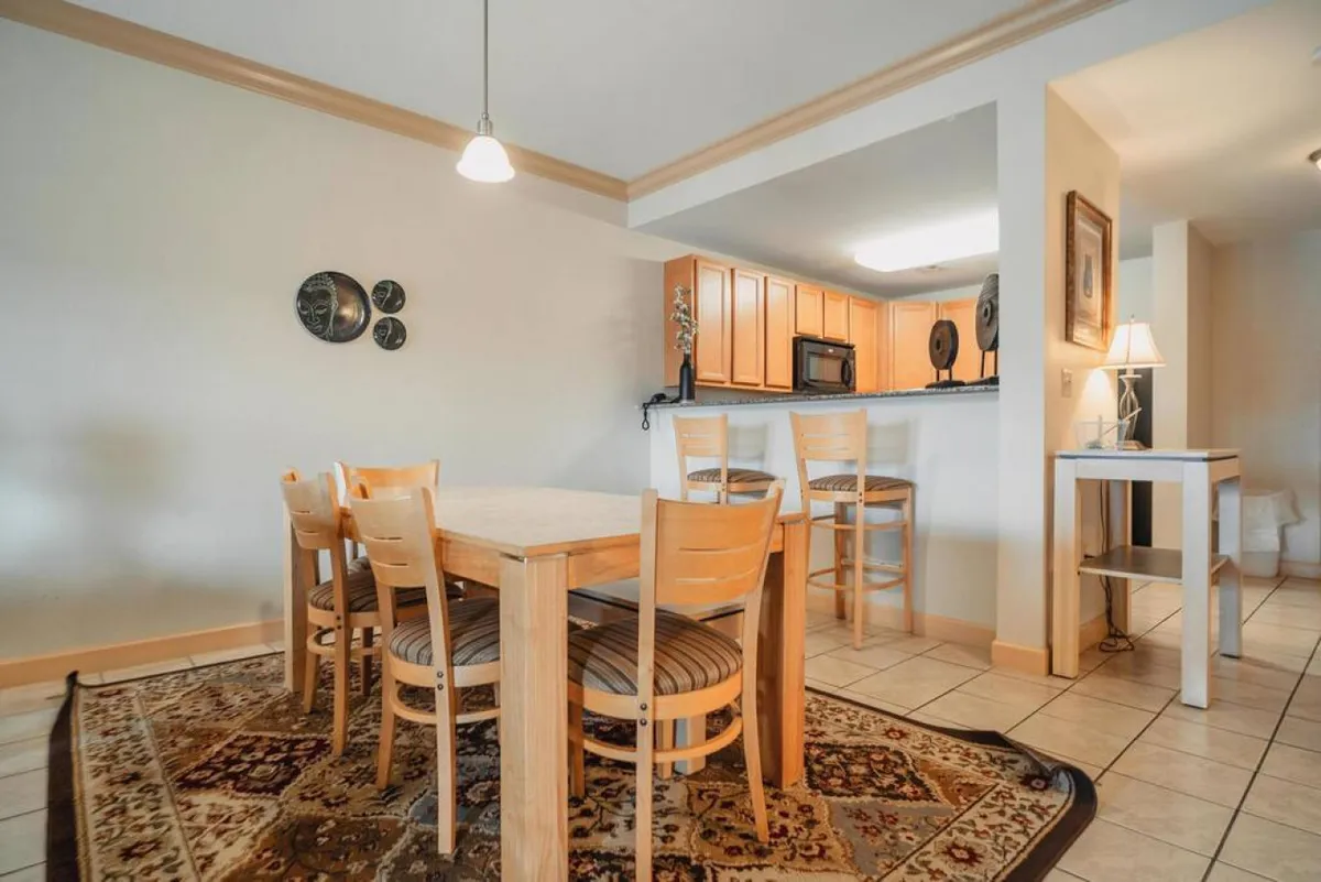 A cozy dining area directly adjacent to the kitchen, with a wooden dining table and chairs set atop a patterned rug. The kitchen counter is visible, offering a glimpse of ample workspace and dishware. Decorative plates on the wall add a festive, homey touch.