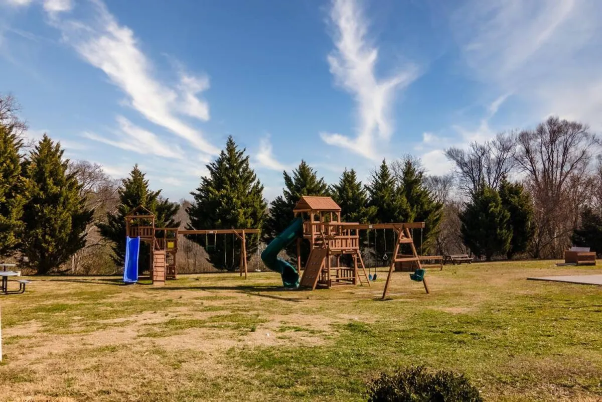 A modern playground structure stands on lush green grass, bordered by trees. Swings, a slide, and open space invite families to gather and children to play, fostering a sense of community and outdoor enjoyment.