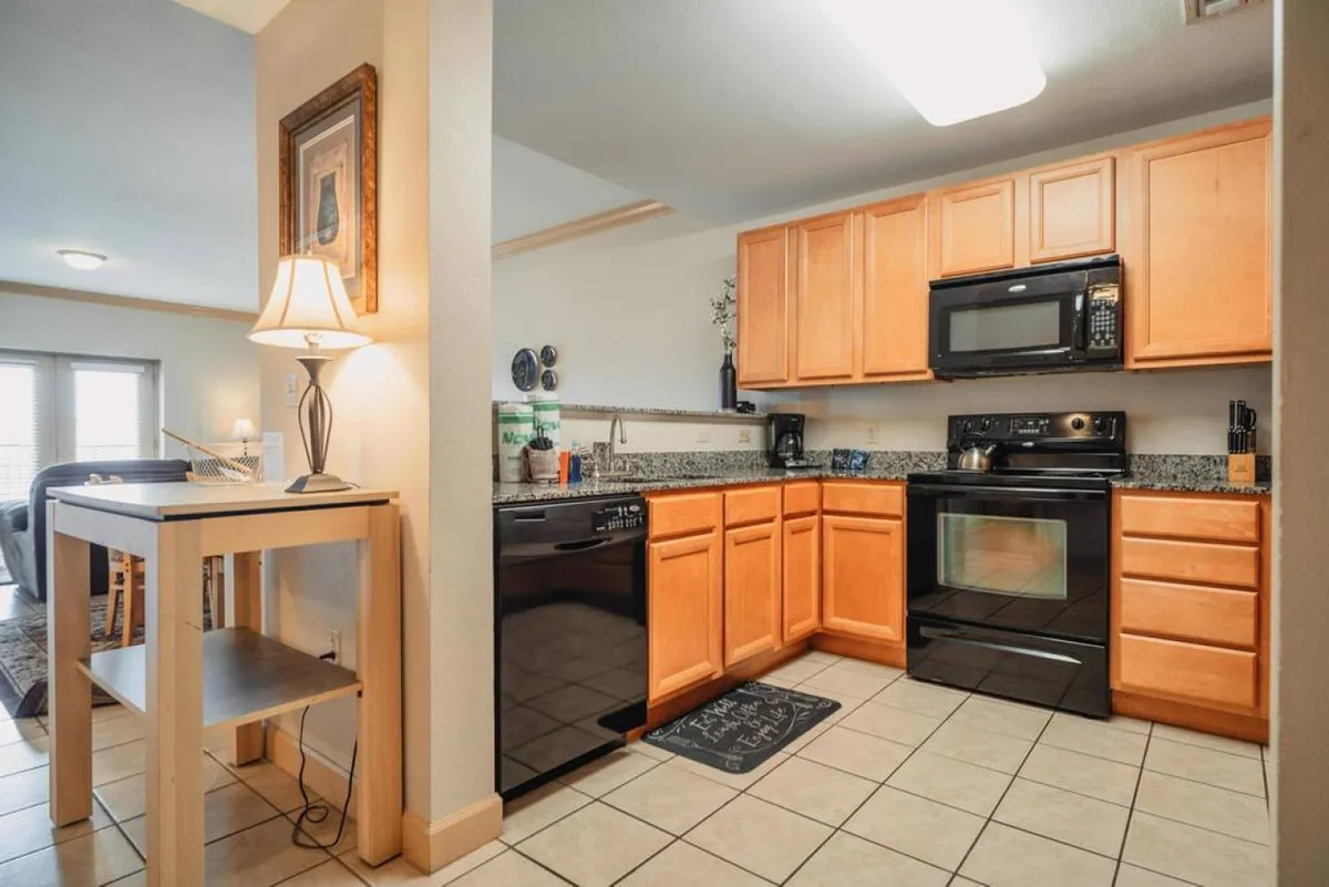 A warm and inviting kitchen and dining area featuring rich wood cabinetry, modern black appliances, a spacious granite countertop, and a cozy dining table illuminated by a table lamp. The open layout connects the kitchen with the dining space, encouraging interaction and socializing while meals are prepared.