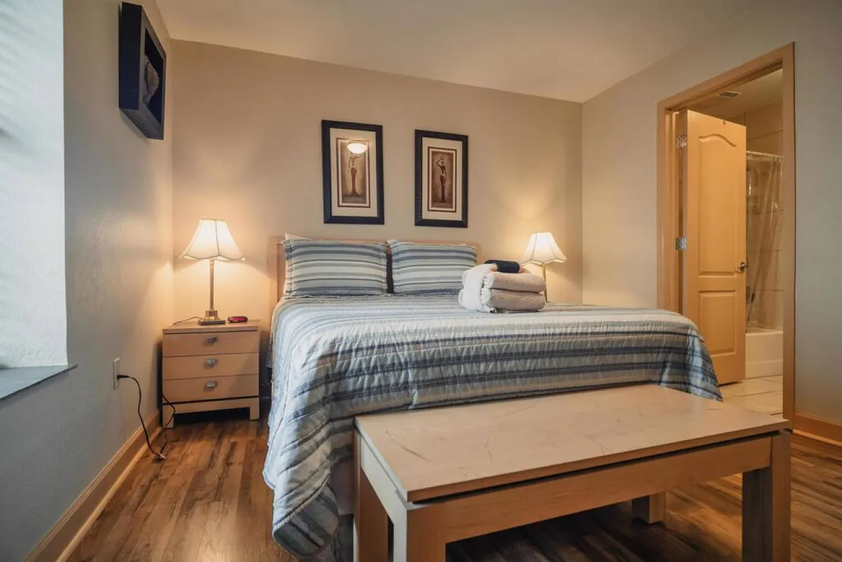A cozy bedroom with a large bed, nightstands, soft lighting from bedside lamps, and tasteful artwork adorning the walls. Gentle natural light filters in, creating an inviting and restful atmosphere.